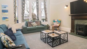 A living room with a sofa, a coffee table, a fireplace, and large windows with curtains. - Rocklands (Penrith)