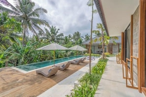 A poolside area with lounge chairs and umbrellas, surrounded by lush greenery and a modern building structure. - Villa The Kayu by GORO (Kabupaten Gianyar)