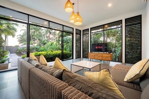 A modern living room with a sofa set, a coffee table, and a TV on a wooden cabinet. Large windows offer a view of greenery outside. - Villa The Kayu by GORO (Kabupaten Gianyar)