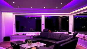 Ein modernes Wohnzimmer mit einer schwarzen Ledersofa, zwei Sesseln und einem Couchtisch. Große Fenster bieten Blick auf die beleuchtete Stadt bei Nacht.