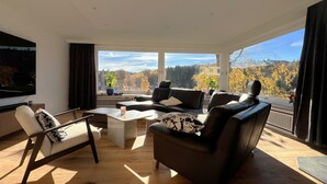 Ein modernes Wohnzimmer mit großem Fernseher, bequemen Sitzmöglichkeiten, einem Couchtisch und Blick auf Herbstbäume durch große Fenster.