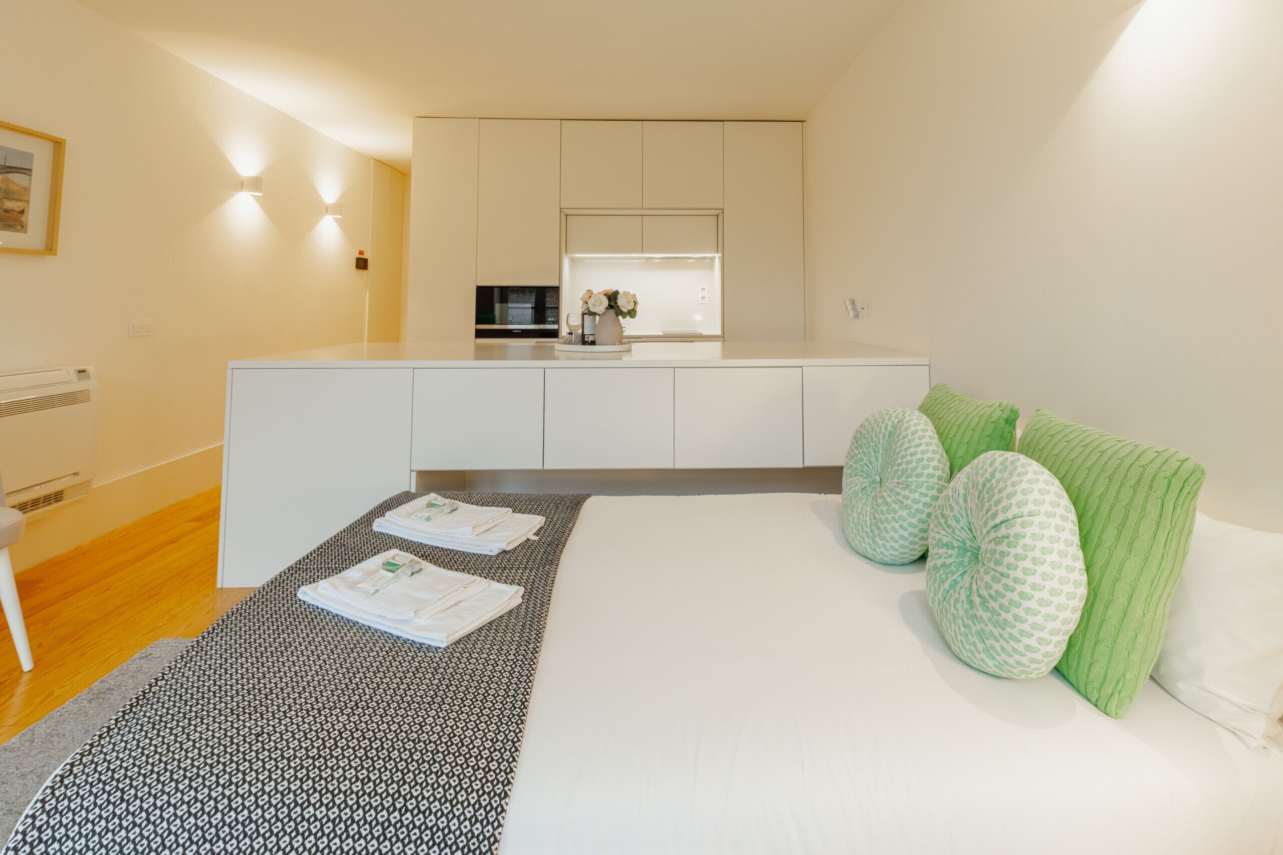 A modern hotel room with a bed, pillows, a bedside table, and a view of the kitchen.