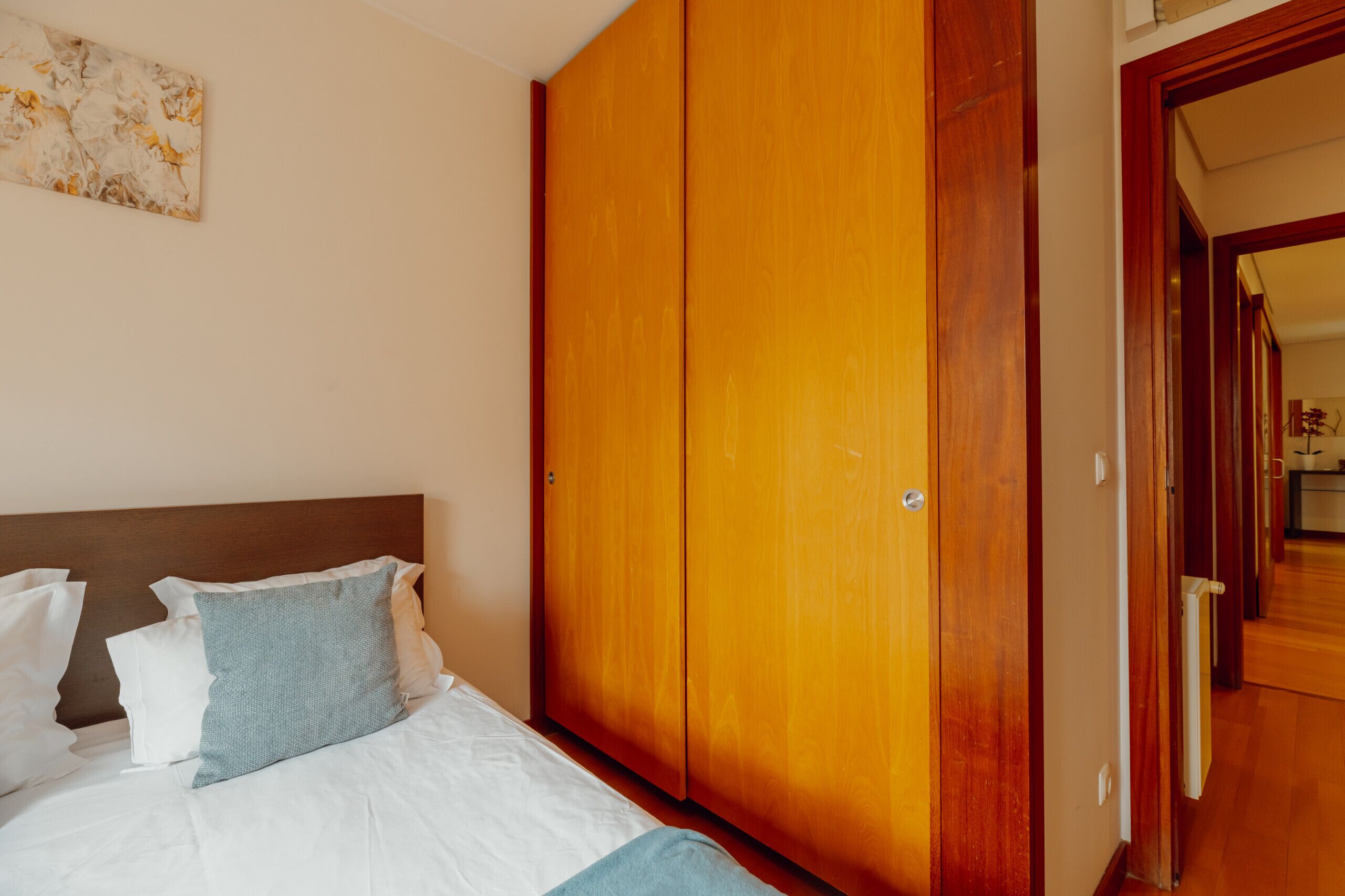 A bedroom with a wooden wardrobe, a bed with white bedding, and a painting on the wall.