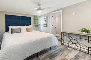 Um quarto com cama, ventilador no teto, janela com persianas, porta e uma pequena mesa com uma planta.