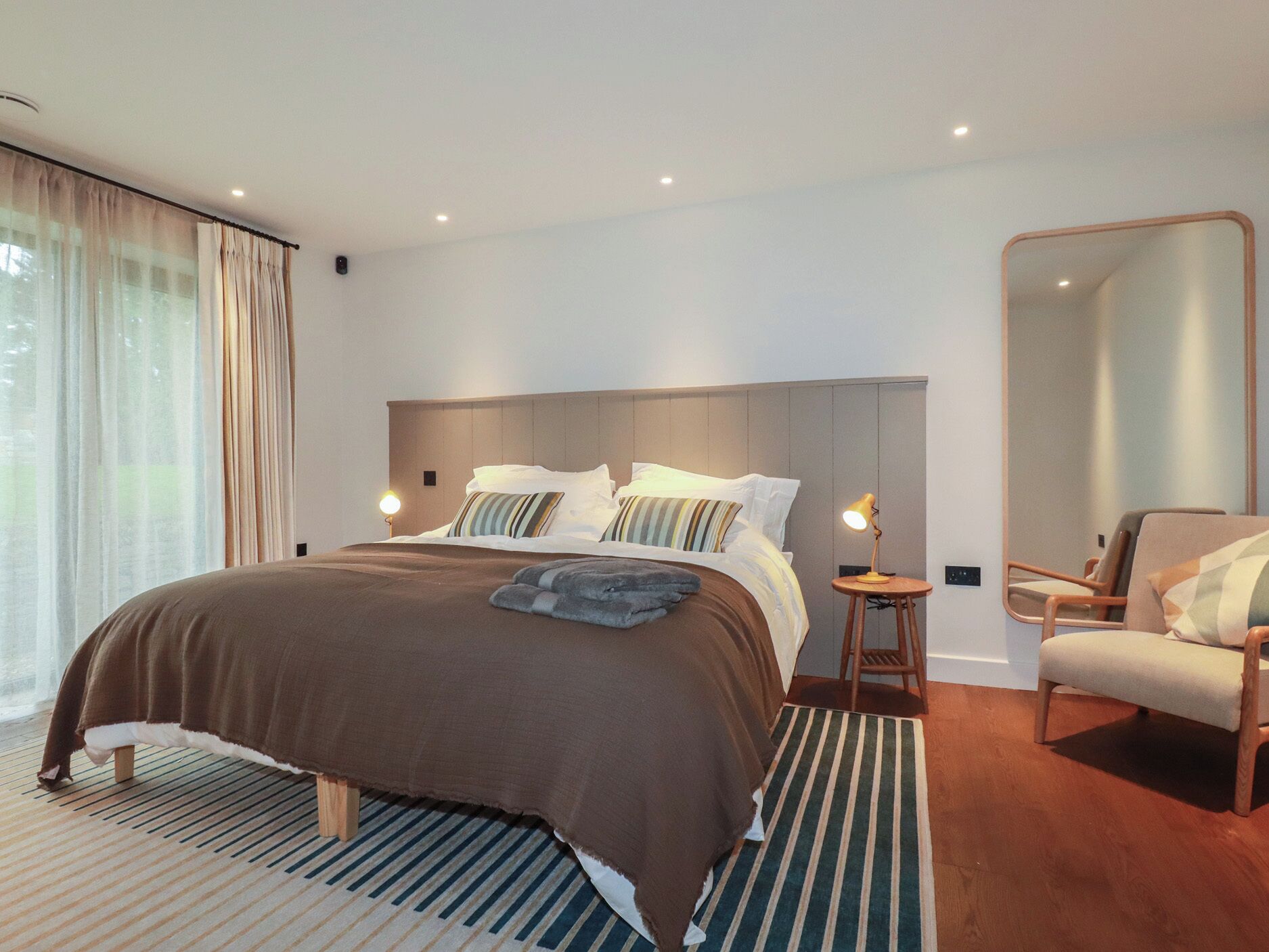 A modern bedroom with a large bed, a bedside table with a lamp, a chair, and a large mirror.
