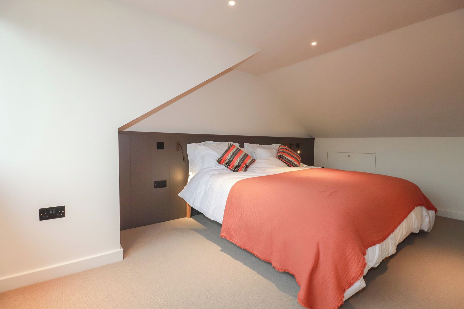 Une chambre avec un plafond en pente, un lit avec une literie blanche et une couverture rouge, deux oreillers à motifs rayés et un luminaire fixé au mur.