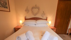 A neatly made bed with white linens and rolled towels, two bedside lamps, and decorative heart-shaped wall hangings. - Thrang View (Ambleside)