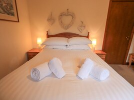 Una cama bien hecha con sábanas blancas y toallas enrolladas, dos lámparas de noche y adornos con forma de corazón en la pared.