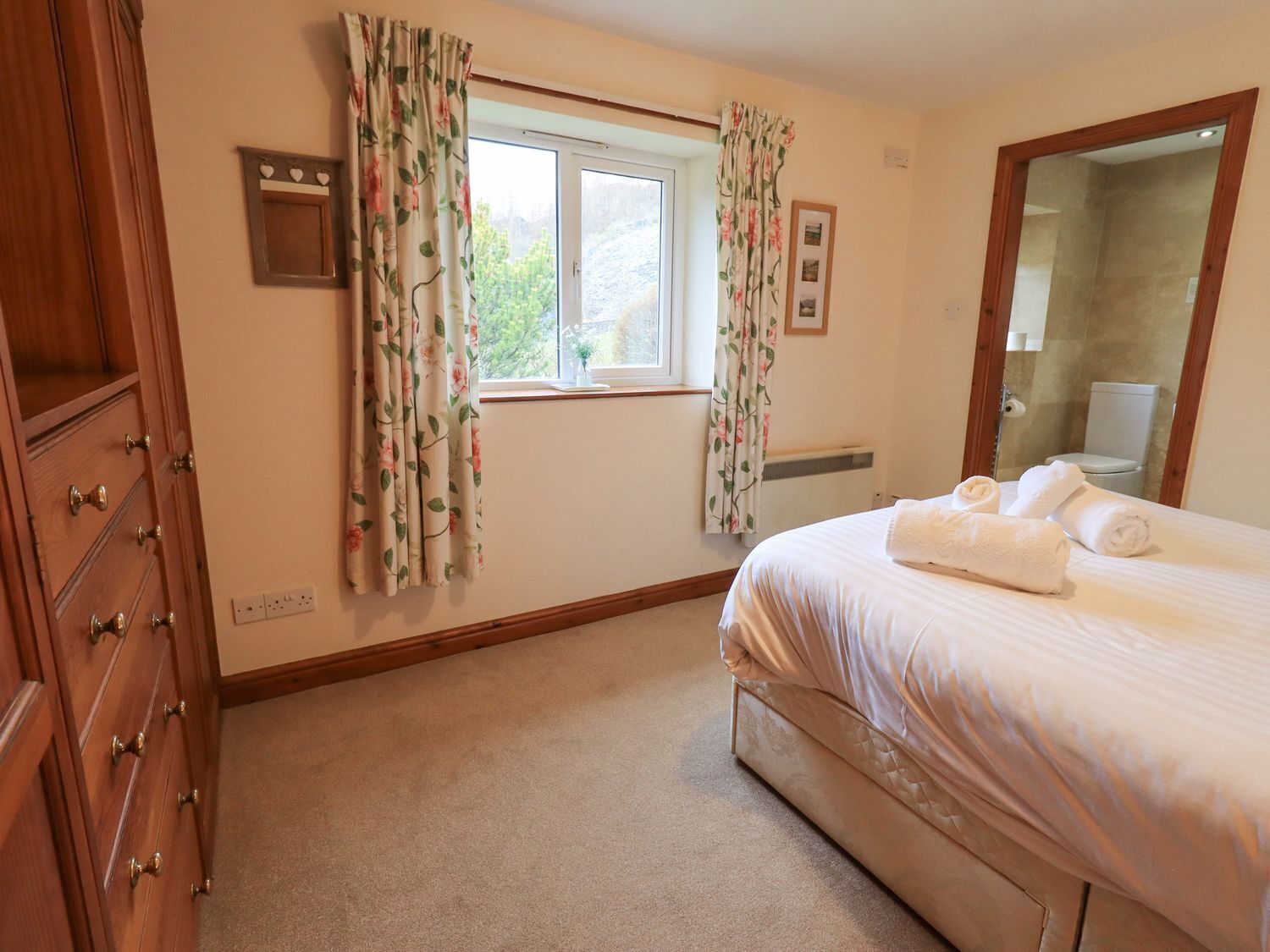 Un dormitorio con cama, una ventana con cortinas florales, un armario de madera y un espejo en la pared.