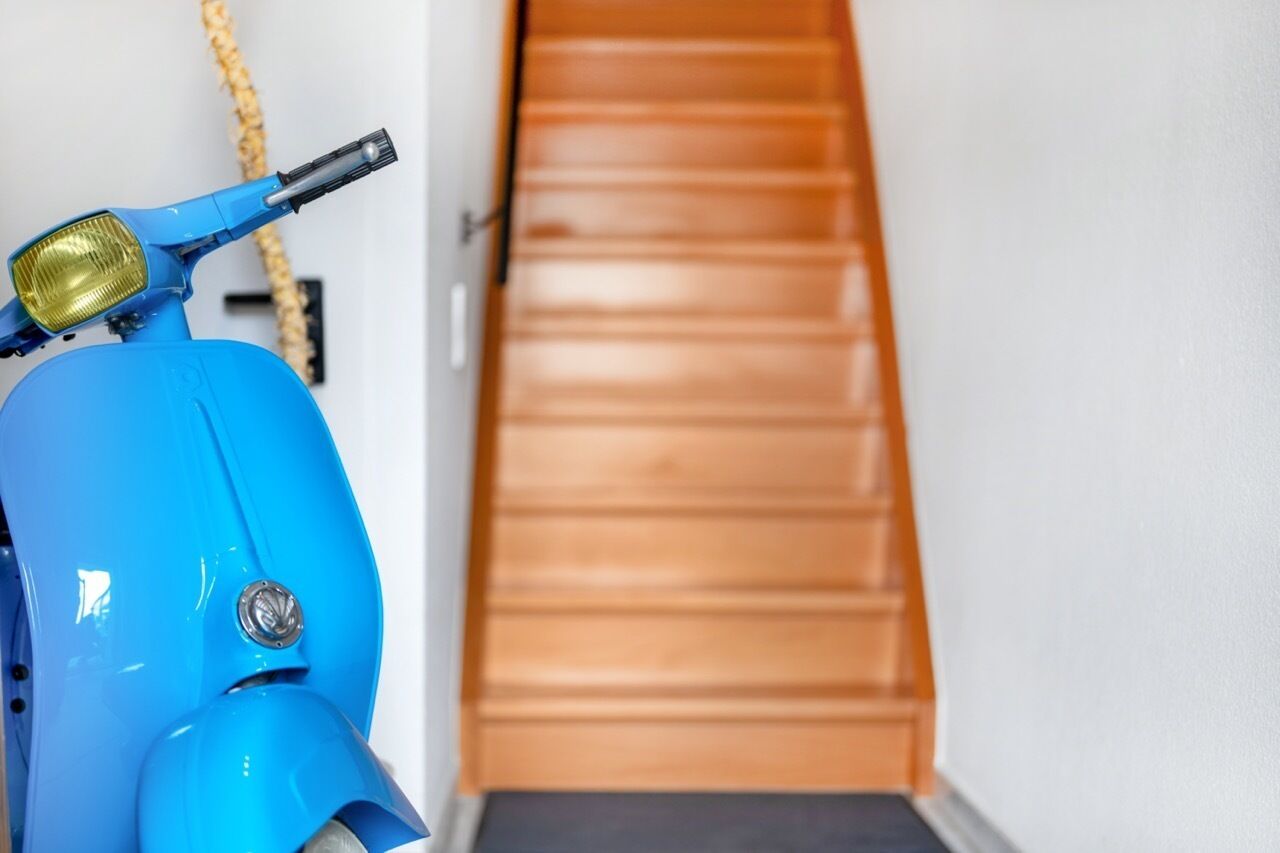 Een blauwe scooter staat binnen met een houten trap op de achtergrond.