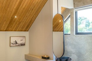 A modern bathroom with a wooden ceiling, a large mirror, a framed picture on the wall, and a window providing natural light. - Villa Bayu by GORO (Bali)