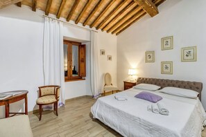 Ein Schlafzimmer mit Holzbalkendecke, einem Bett mit weißer Bettwäsche, einem Sessel, einem Tisch und einem Fenster mit Vorhängen.