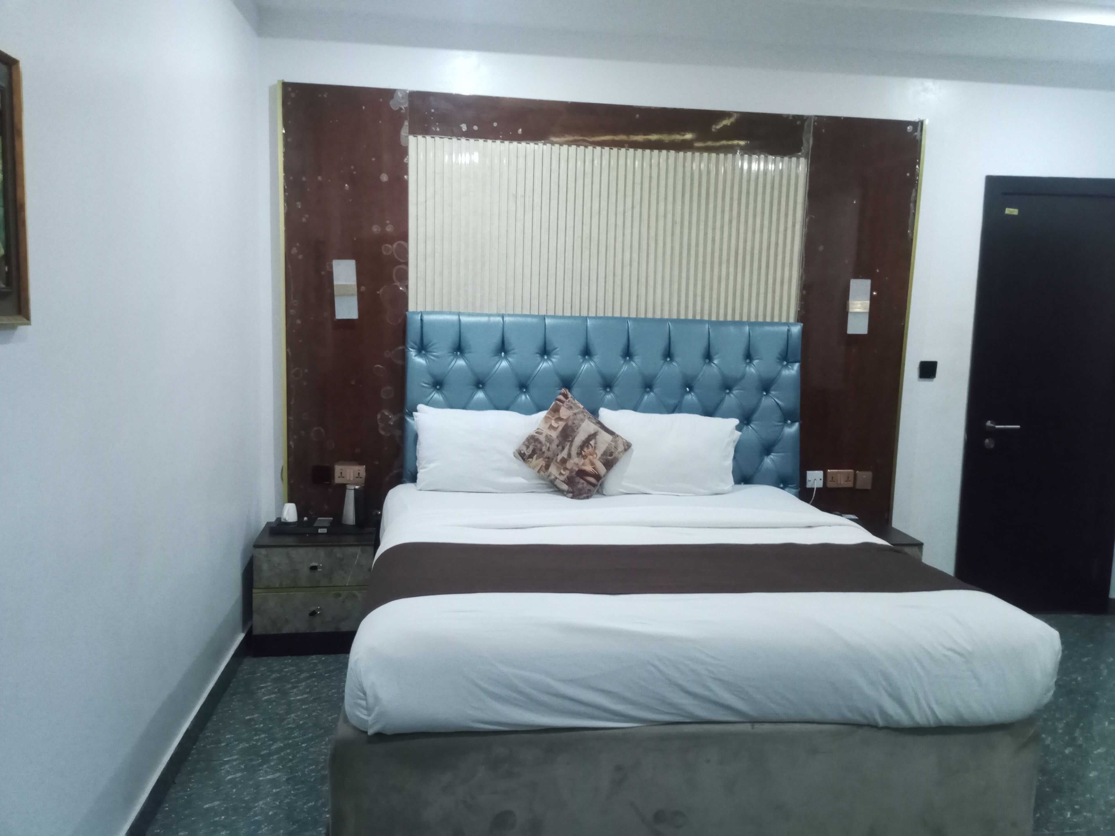 A hotel room with a large bed, a nightstand, a black door, and a window with blinds.