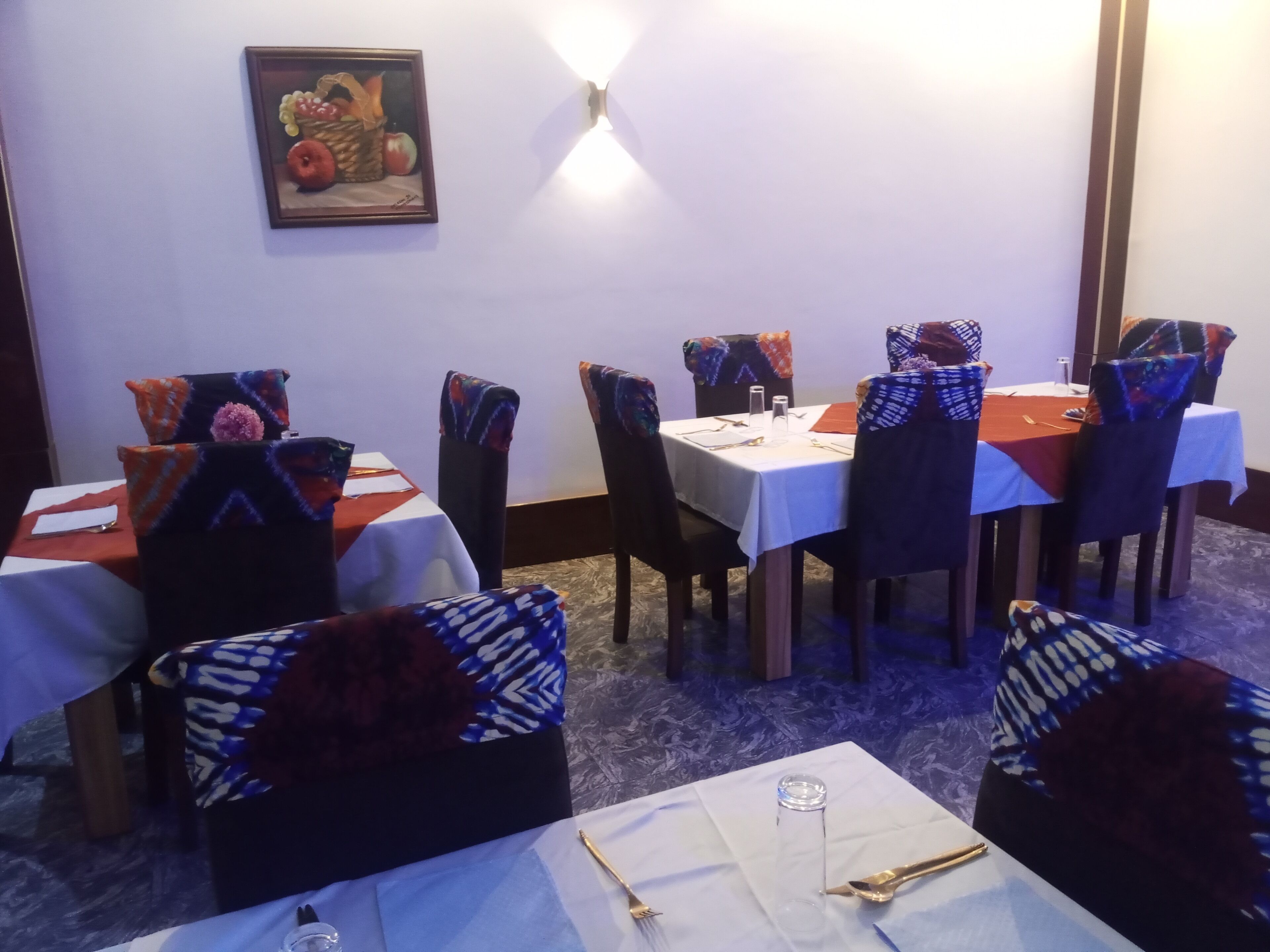 A dining area with tables set for four, chairs with patterned covers, and a wall-mounted light fixture.
