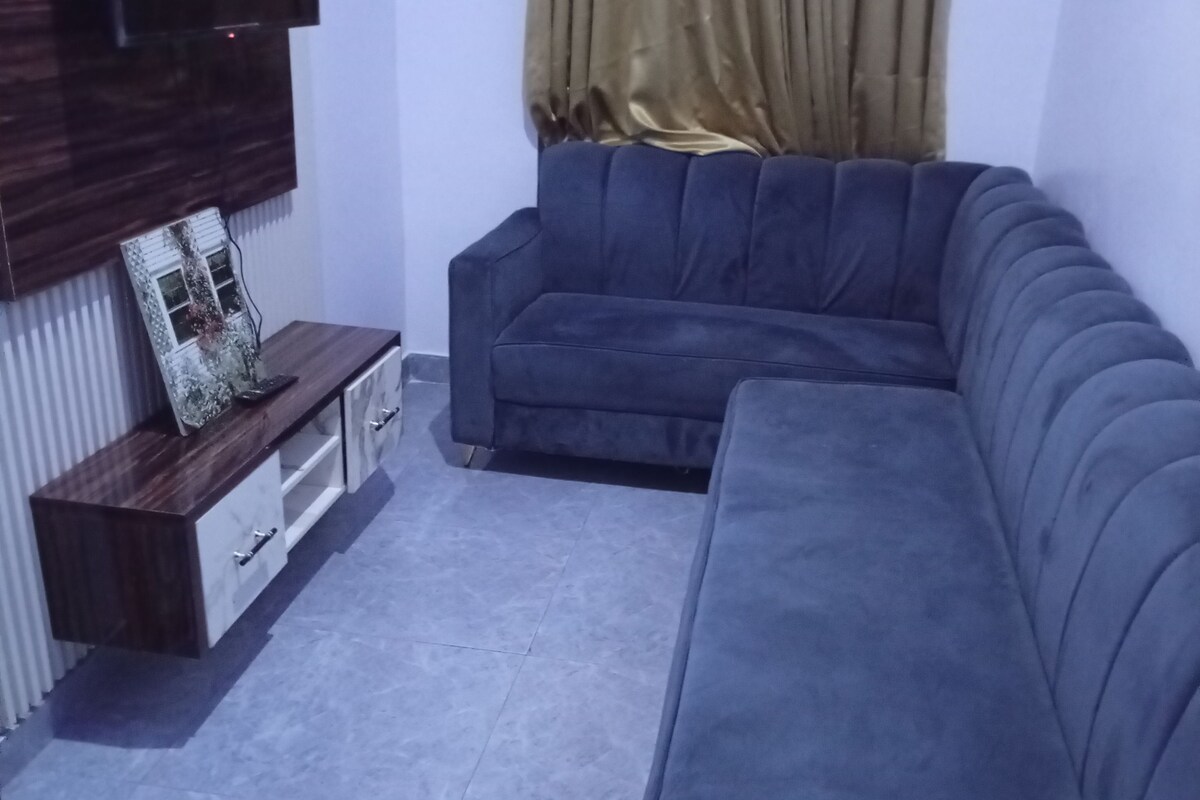 Een moderne woonkamer met een grijze modulaire bank, een houten tv-meubel en een groot raam met gordijn.