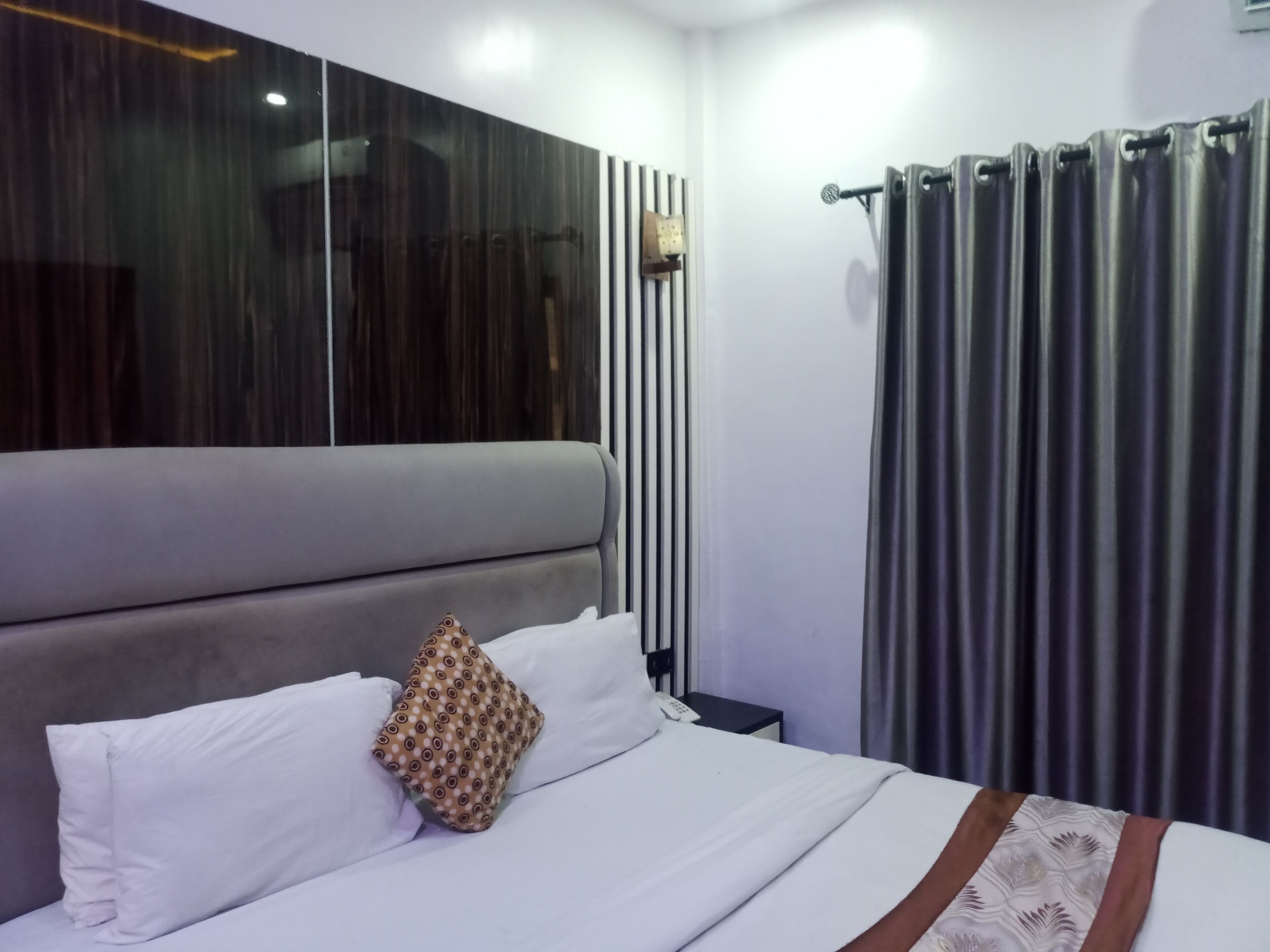 A modern hotel room with a large bed, a headboard with vertical lines, a bedside table with a lamp, and a curtain on the window.