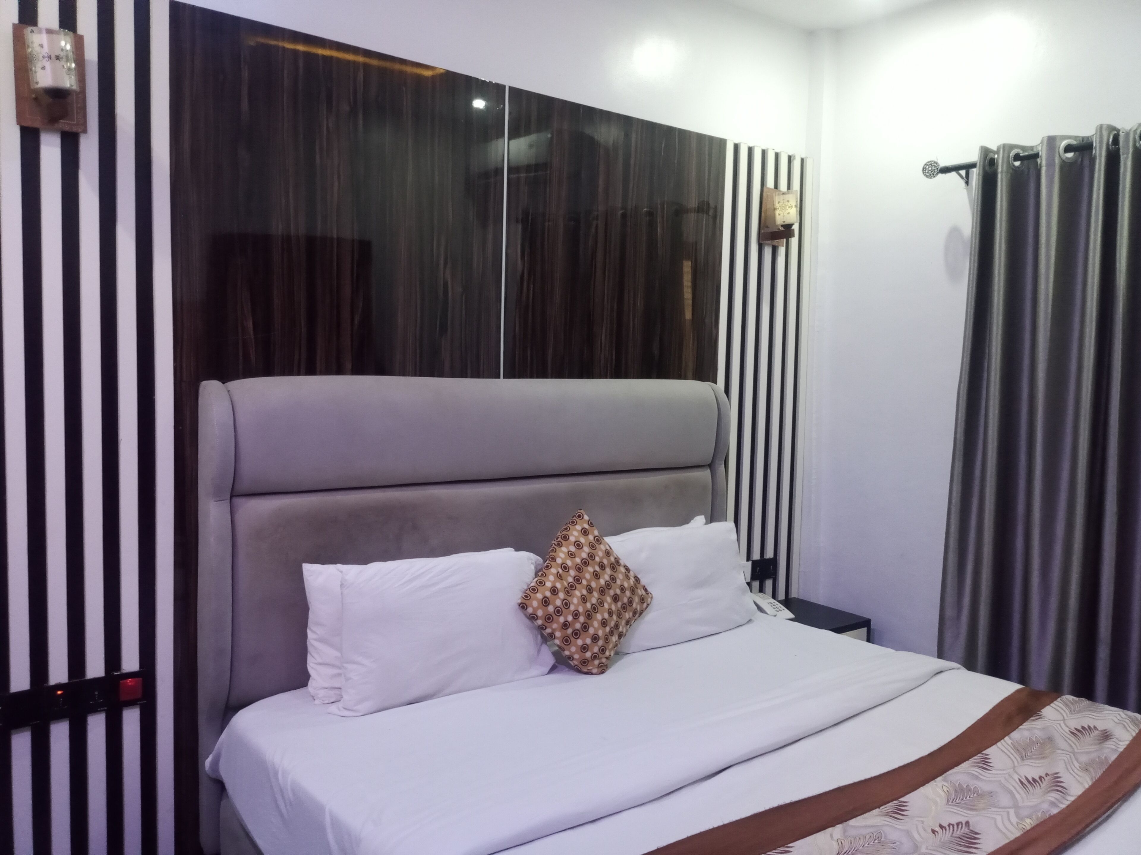 Una cama bien hecha con sábanas blancas y un cojín estampado, un cabecero tapizado en gris y una pared con rayas verticales negras y blancas.