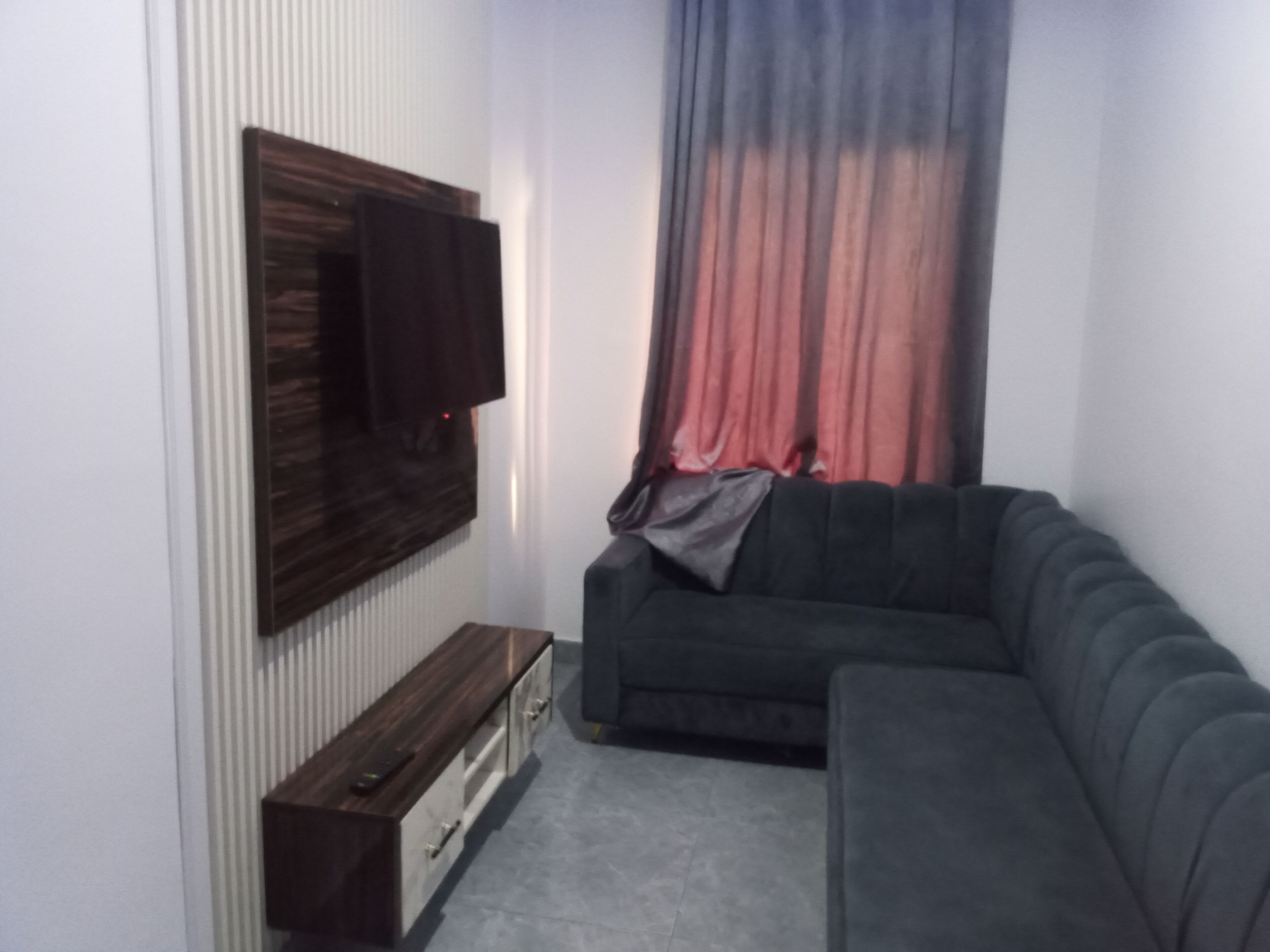 A modern living room with a sectional sofa, a flat-screen TV mounted on the wall, a wooden cabinet, and a red and blue curtain.