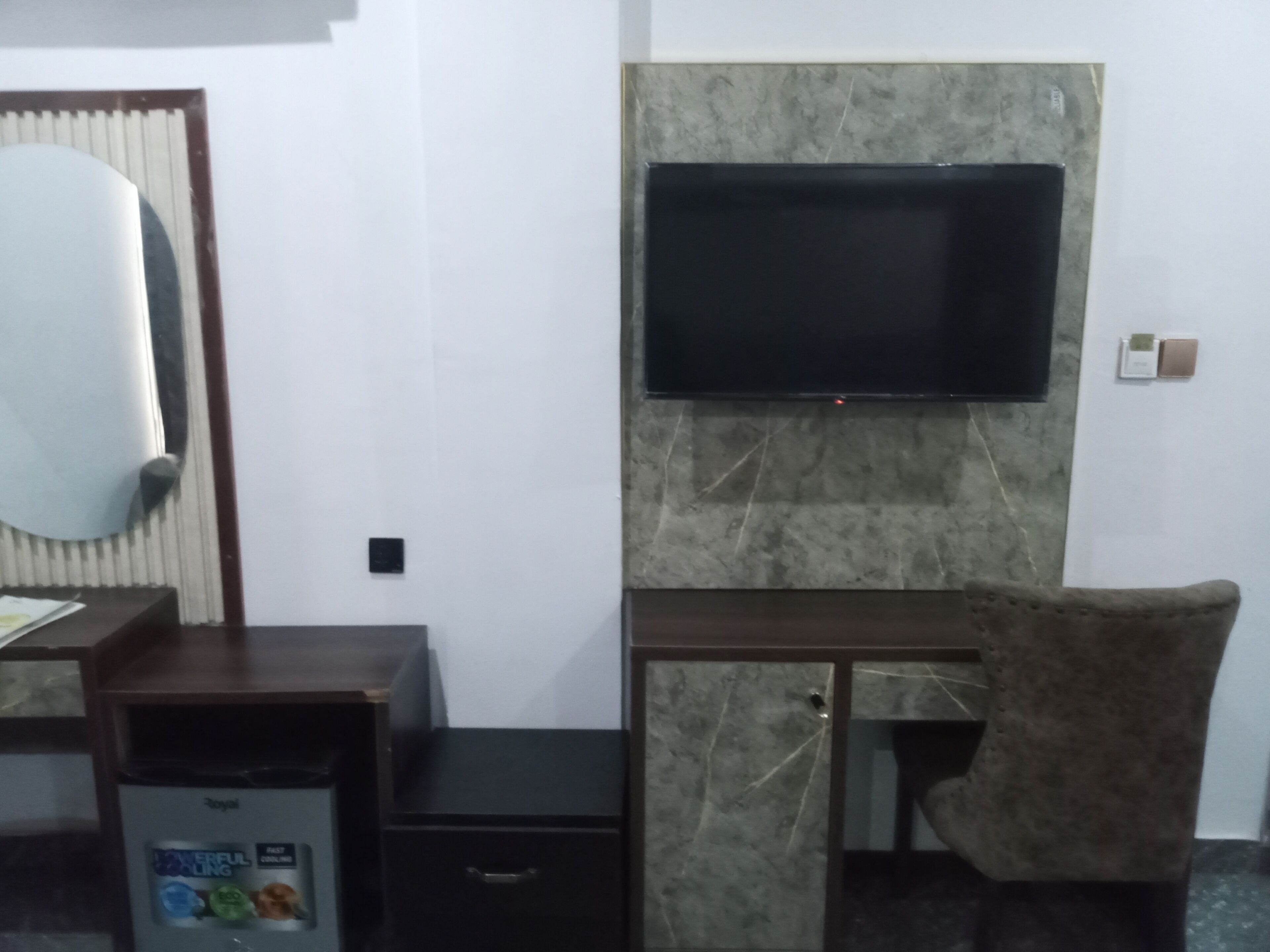 A room with a flat-screen TV mounted on a marble wall, a wooden desk with a chair, and a small refrigerator.