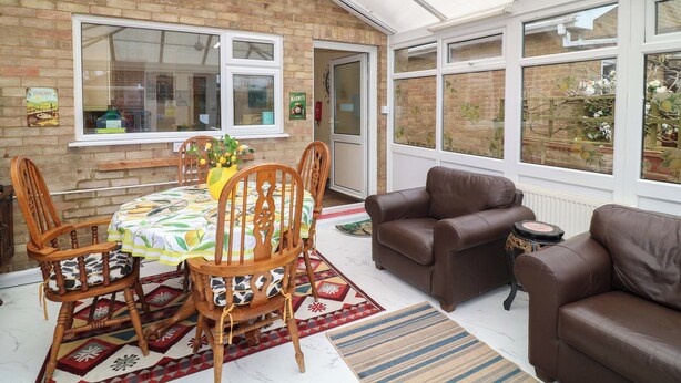 A conservatory with a brick wall, a dining table with chairs, a sofa, and a small table with a record player. - 5 Overstone Drive (Wisbech)