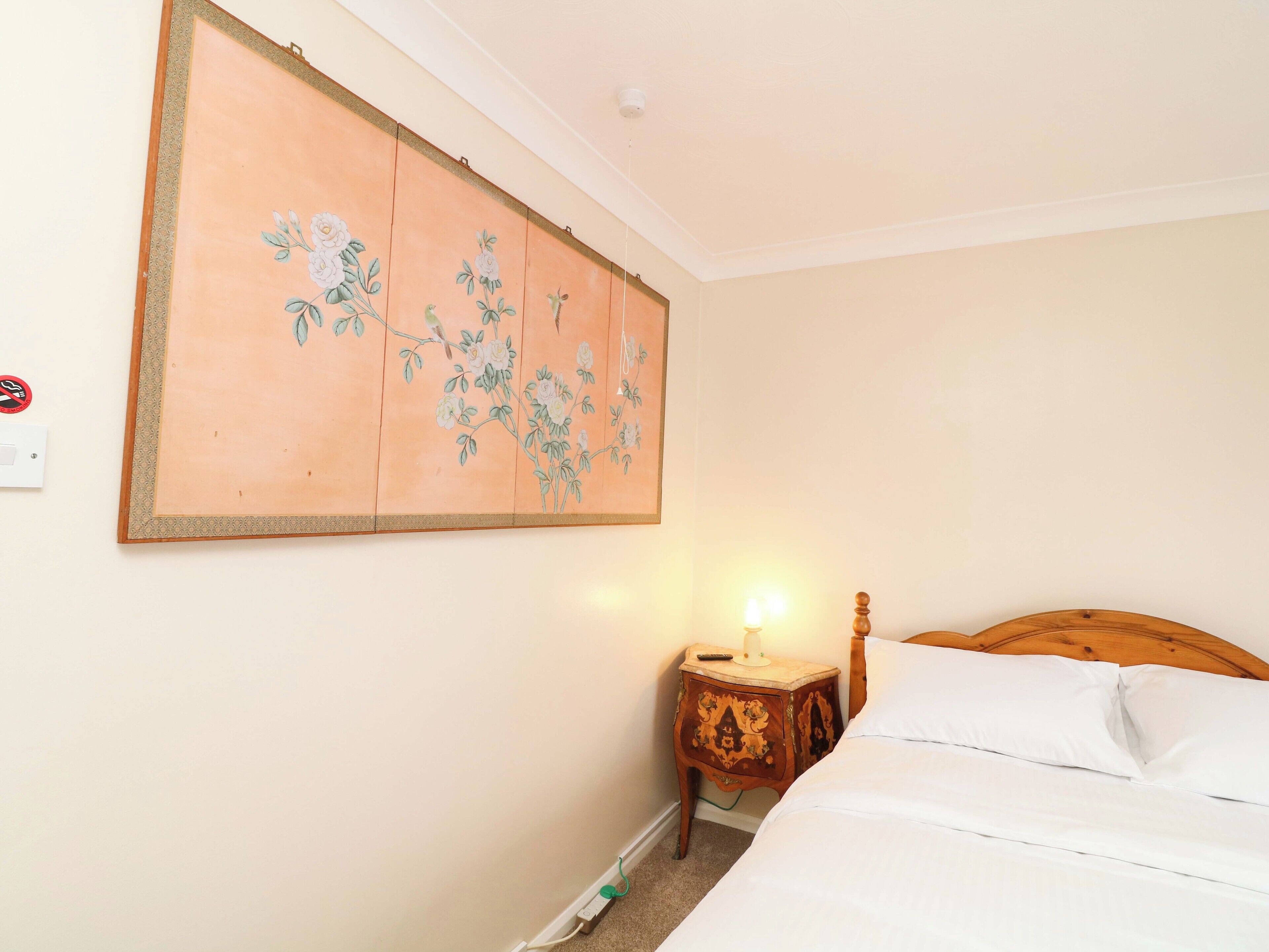 A bedroom with a bed, a nightstand with a lamp, a wall-mounted painting, and a power outlet.