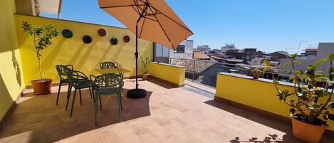 A rooftop terrace with yellow walls, a table with chairs, a potted plant, and a large umbrella.