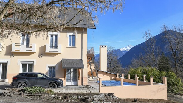 Un edificio di due piani con un balcone, un'auto parcheggiata davanti e un paesaggio montano sullo sfondo.