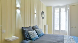 Una camera da letto con un letto, cuscini, un comodino, una lampada a parete, un termosifone e una finestra vista.