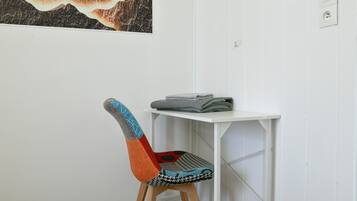 Une chaise avec un dossier coloré et un siège à motifs, une table blanche sur laquelle sont pliés des objets, et un mur orné d’une œuvre d’art encadrée.