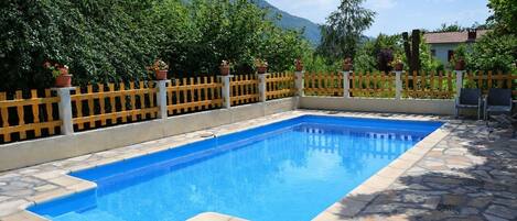 A rectangular swimming pool with a stone border, surrounded by a wooden fence and potted plants.