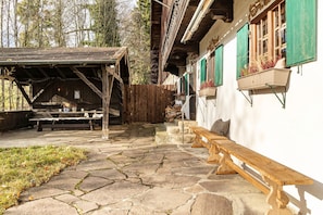A rustic outdoor seating area with stone flooring, wooden benches, and a covered shelter.