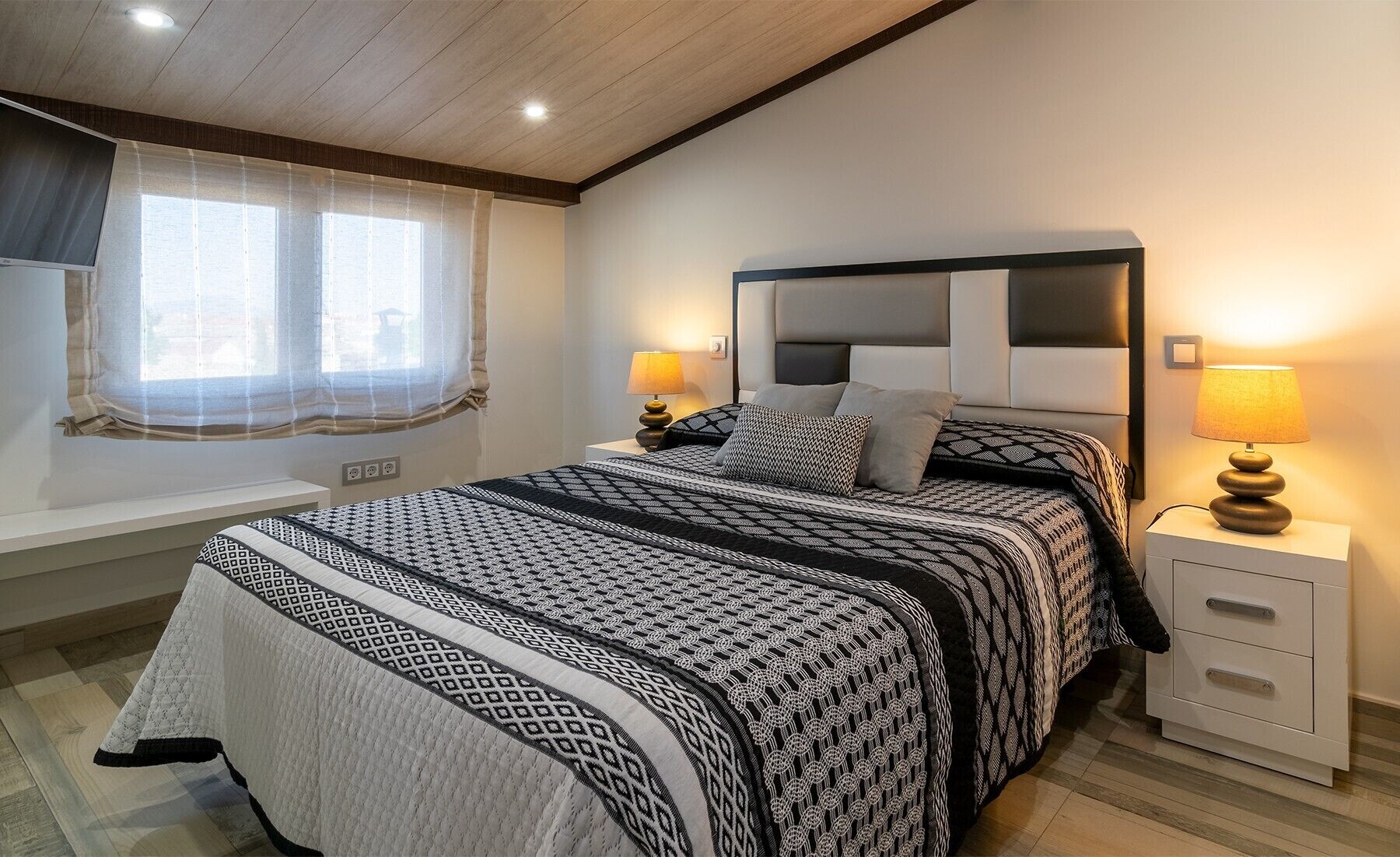 Un dormitorio con cama, mesitas de noche, lámparas, una ventana con cortinas y techo de madera.