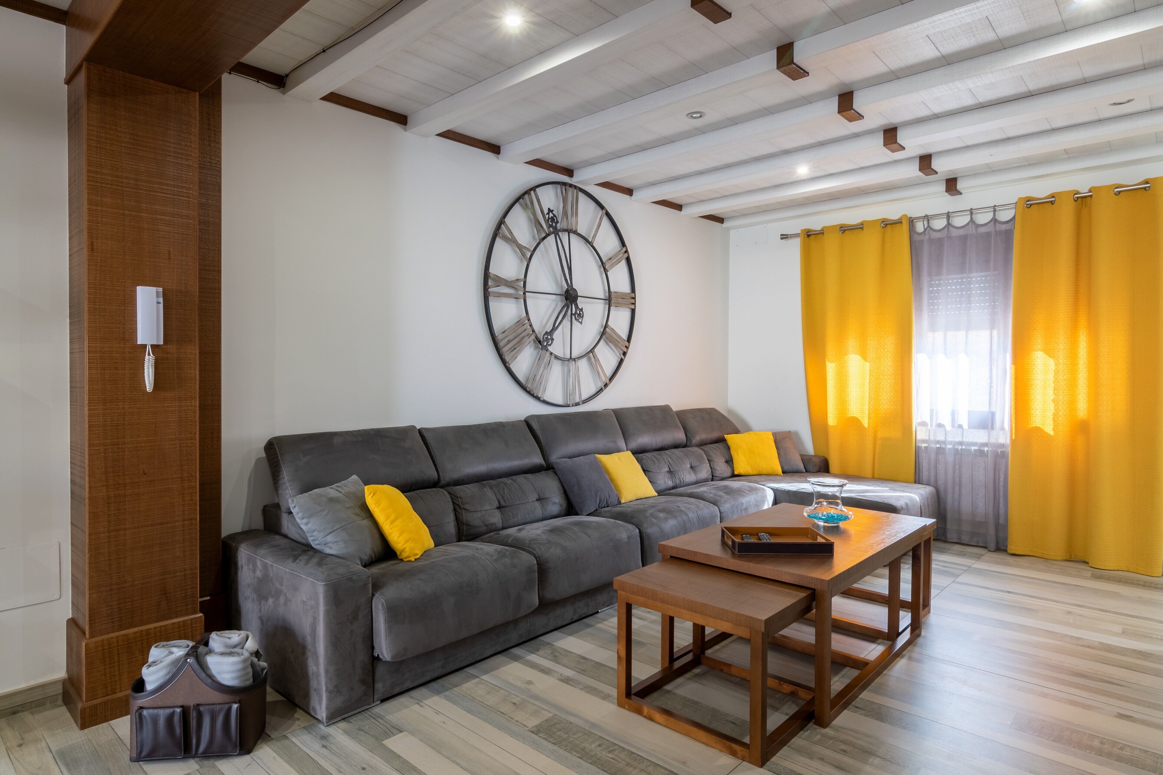 Un salon avec un canapé d’angle, une table basse en bois, une horloge murale et des rideaux jaunes.