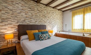 Un dormitorio con pared de piedra, una cama con almohadas estampadas, lámparas de noche y una bañera.
