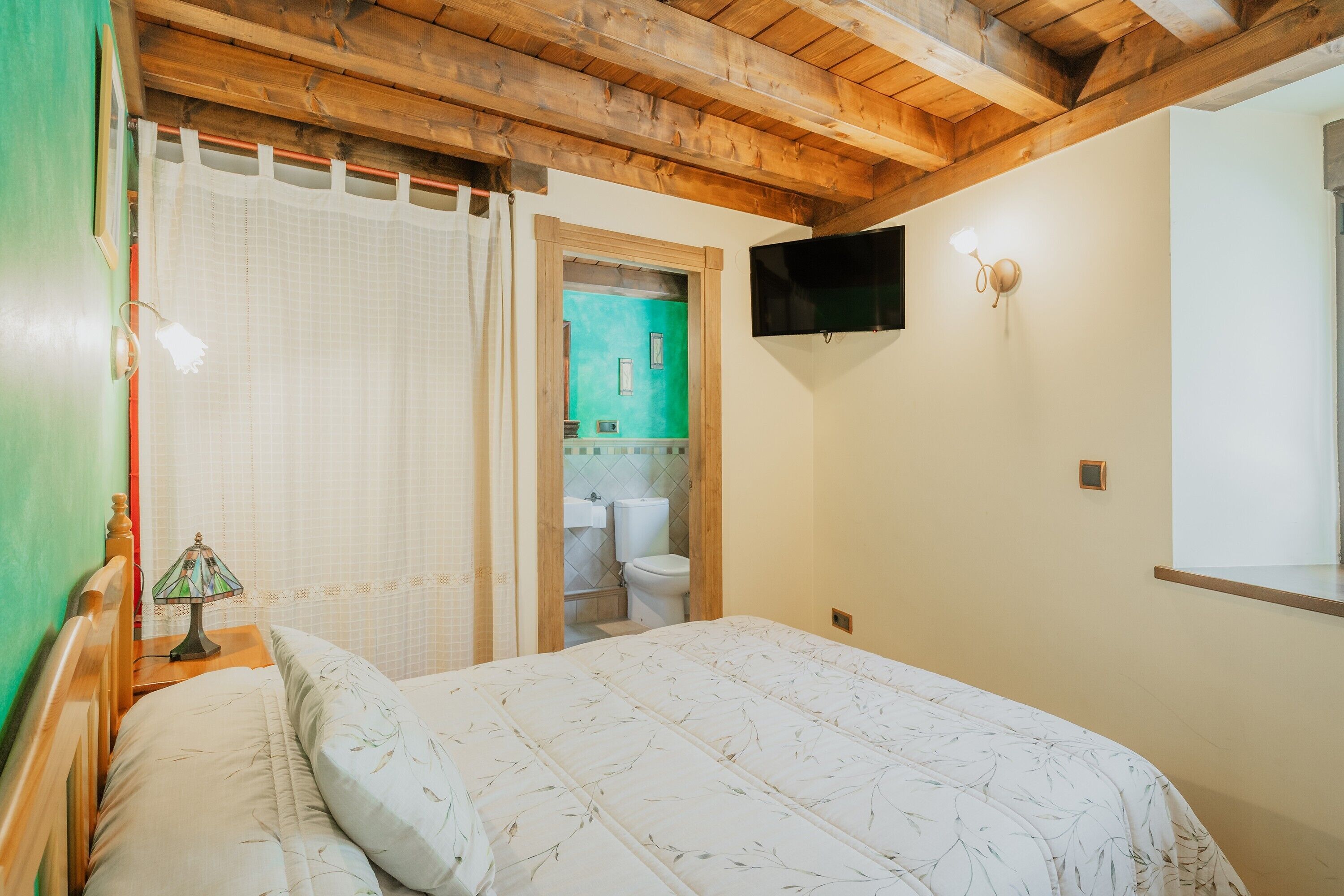 Un dormitorio con techo de madera, una cama con colcha estampada, una lámpara de noche, una pared verde y un televisor montado.