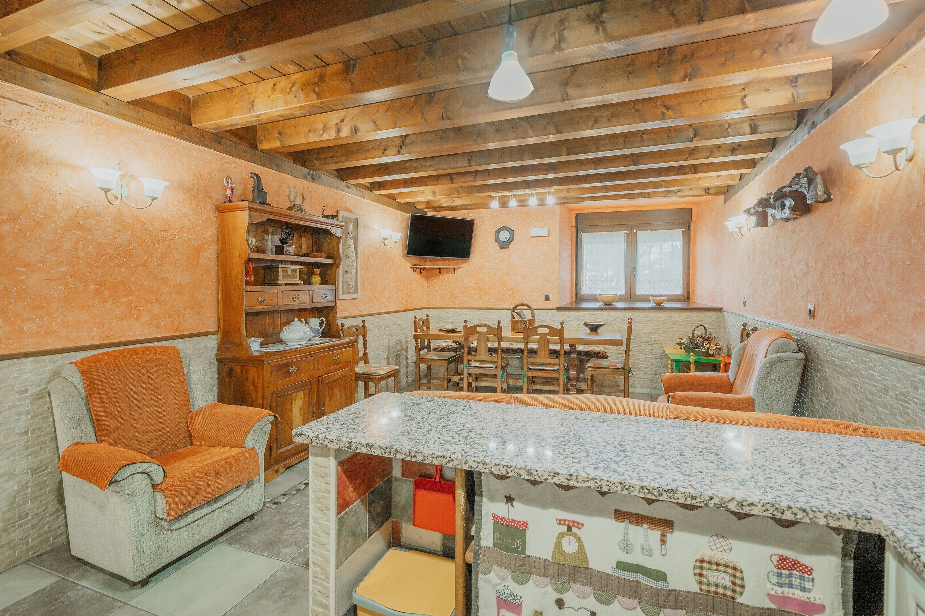 Una habitación acogedora con techo de madera, un televisor instalado en la pared, una mesa de comedor con sillas y una pequeña cocina con mostrador.
