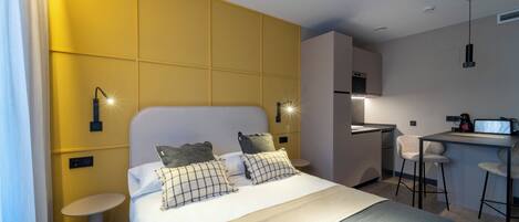 A modern hotel room with a large bed, a small round table, a kitchenette, and a bar area.