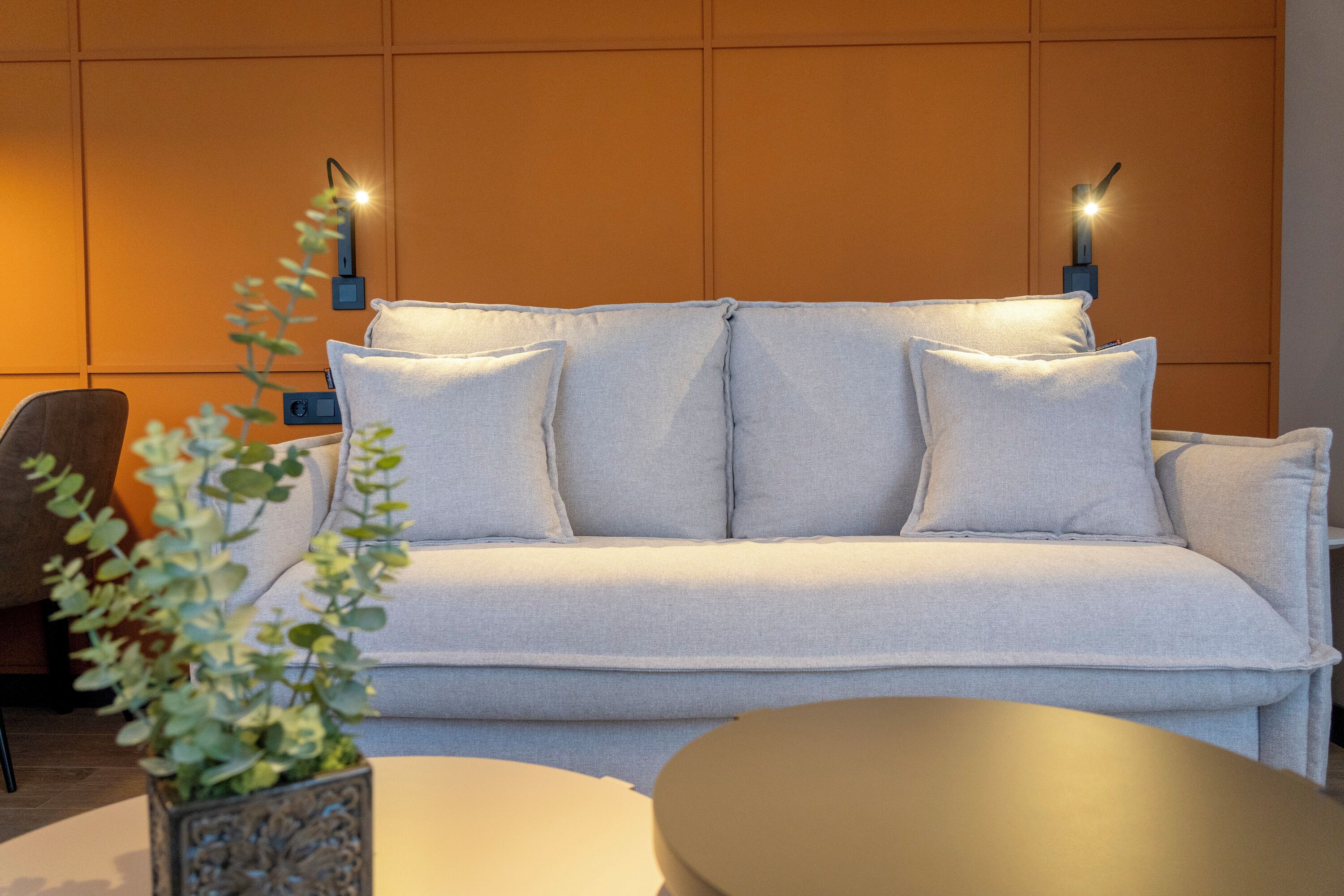Un salon moderne avec un canapé gris clair, deux lampes fixées au mur et une plante décorative.