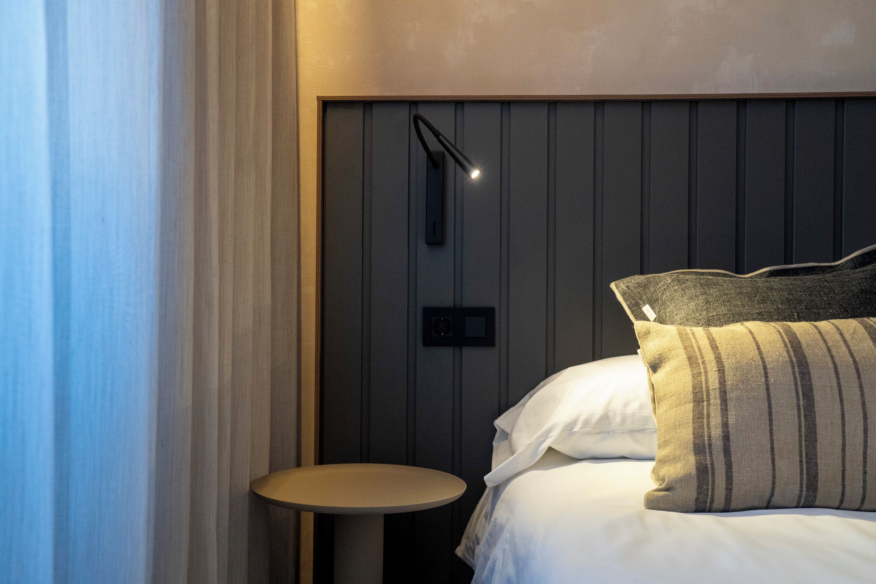 Una cama bien hecha con almohadas blancas y a rayas, una mesita redonda y una lámpara fijada a la pared.