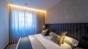 Una habitación de hotel moderna con una cama grande, un cabecero con líneas verticales y un amplio ventanal con cortinas ligeras.