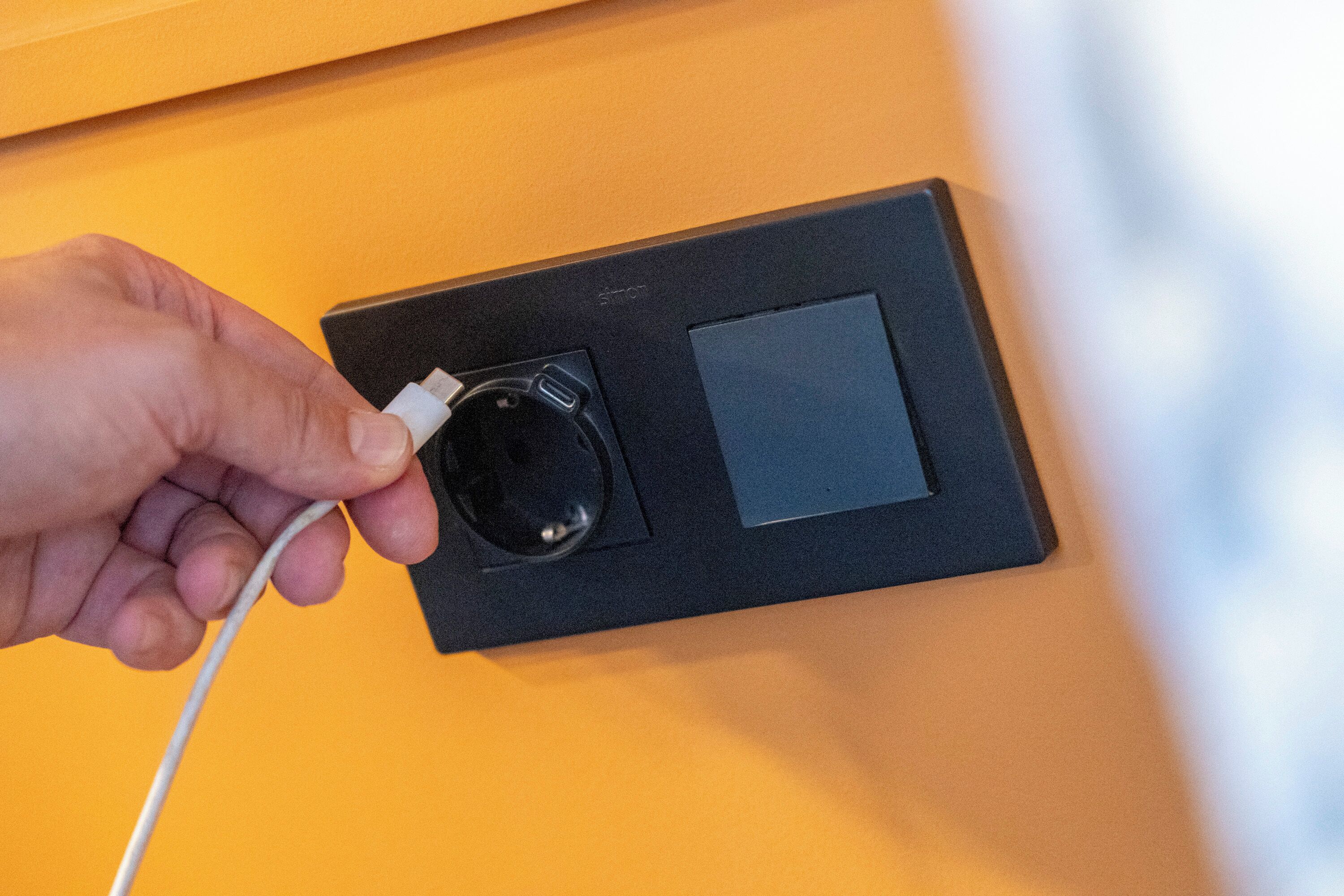 A hand plugging a white USB cable into a black charging port on a yellow wall.