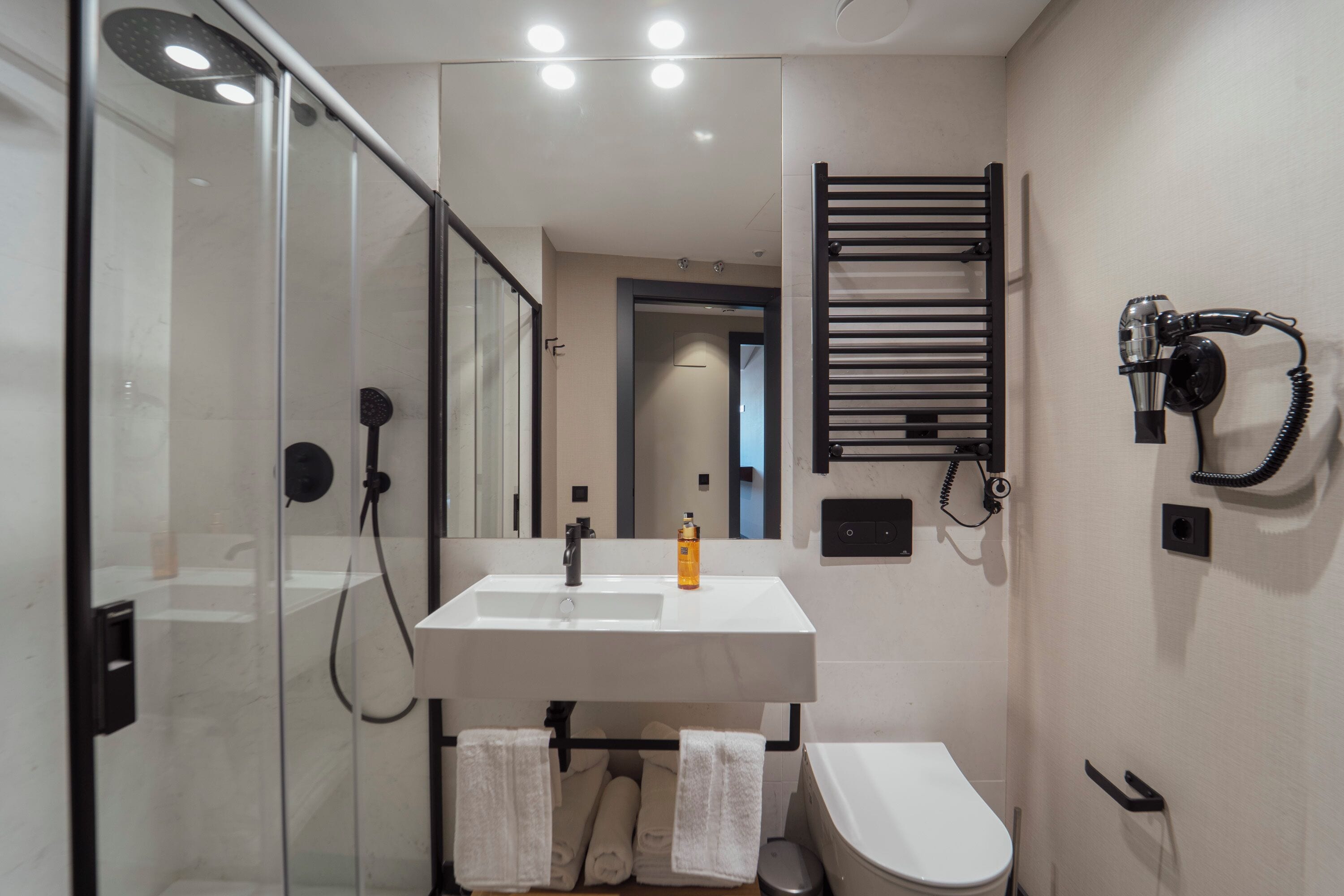 A modern bathroom with a glass shower enclosure, a white sink with a mirror above, a towel rack, and a hairdryer mounted on the wall.