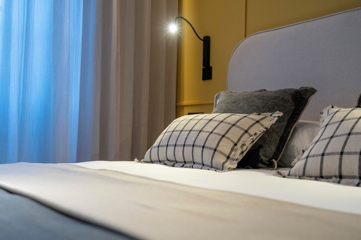 A neatly made bed with a white comforter and two pillows, a wall-mounted lamp, and a window with curtains.