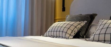 A neatly made bed with a white comforter and two pillows, a wall-mounted lamp, and a window with curtains.