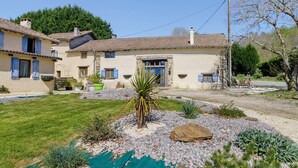 A two-story house with a stone facade, a front entrance with a door, and a small garden with a palm tree and rocks. - Country House “Le Ranch Des Ours” with Private Pool, Terrace, and Wi-Fi (Champsac)