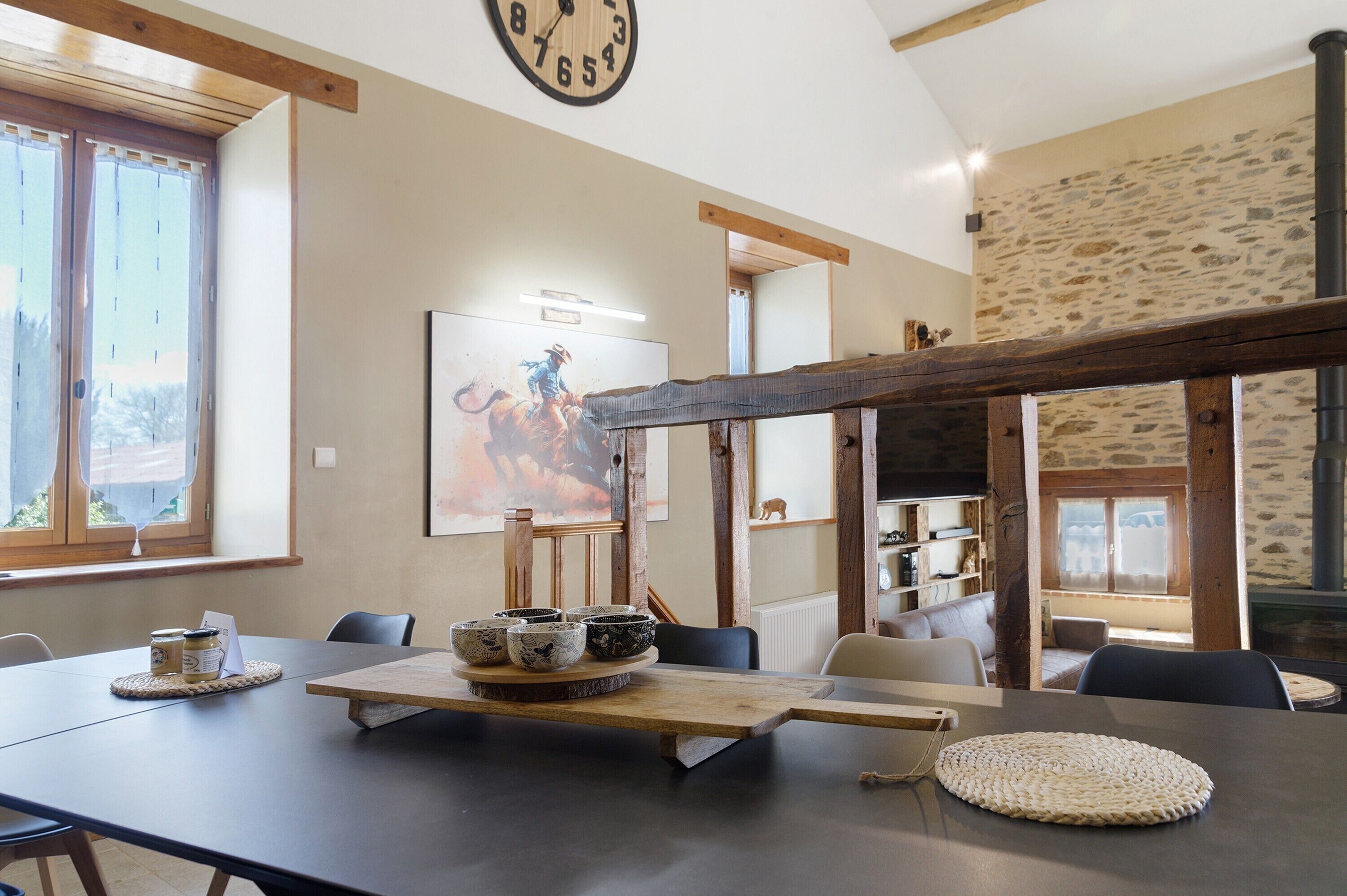 Un comedor con una mesa larga, sillas, techo de vigas de madera, un reloj y una pared de piedra rústica.
