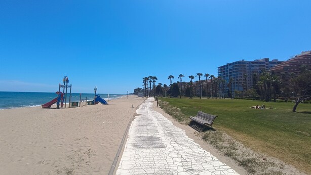 Une plage avec une aire de jeux, une promenade dallée et un espace herbeux agrémenté d’un banc.