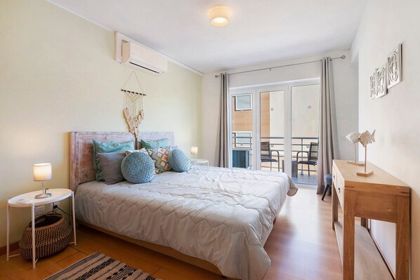 A bedroom with a bed, bedside table, lamp, air conditioning unit, and a view of the balcony.