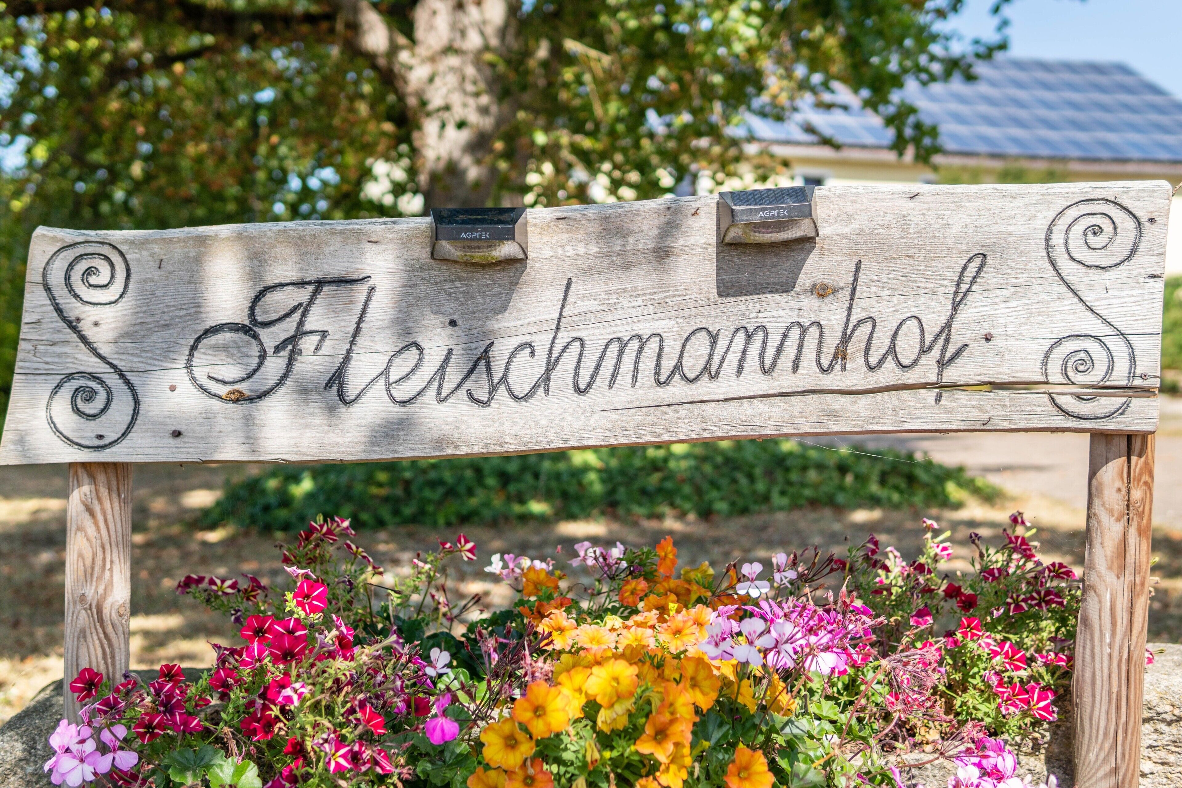 A wooden sign with the word 'Fleischmannhof' written on it, supported by two wooden posts, with a variety of colorful flowers in front.