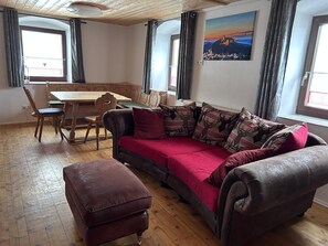 A room with a wooden dining table, a sofa with patterned cushions, a red ottoman, and a painting of a landscape on the wall.